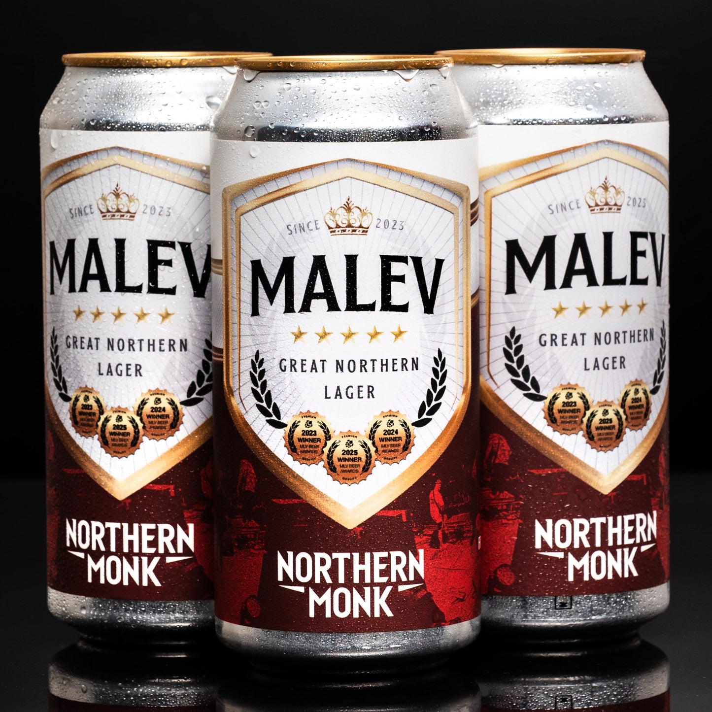 Three cans of MALEV Northern Monk Lager on a reflective surface with a dark background