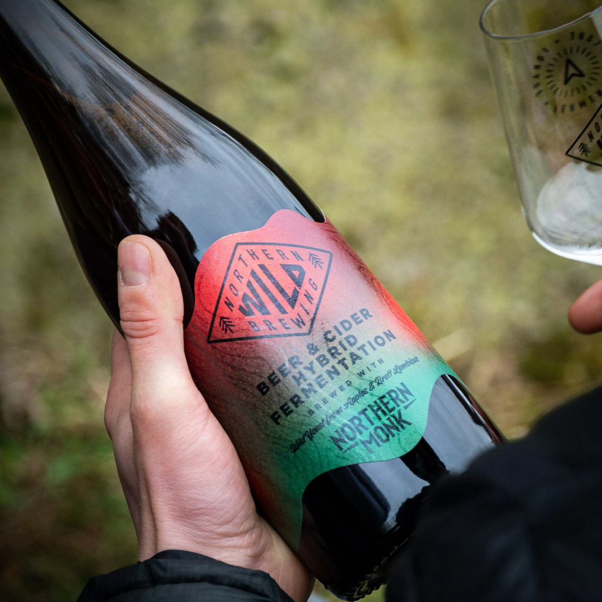 Close-up of a hand holding a bottle of Northern Wild 006 Hogans Cider with colorful label and glass in background.