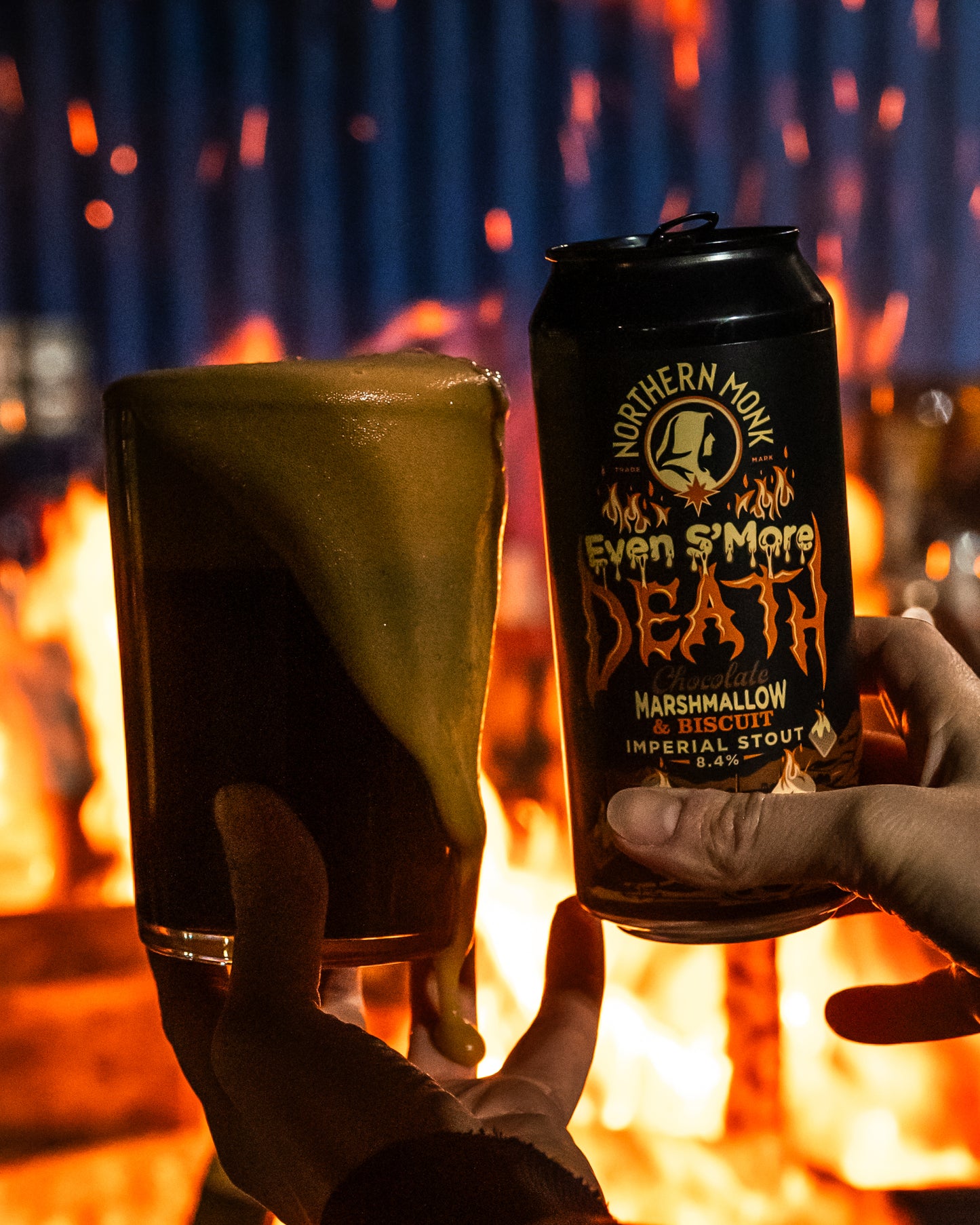 Two hands holding a glass of dark beer and a can of 'Even S'more Death' Imperial Stout in front of a fire.