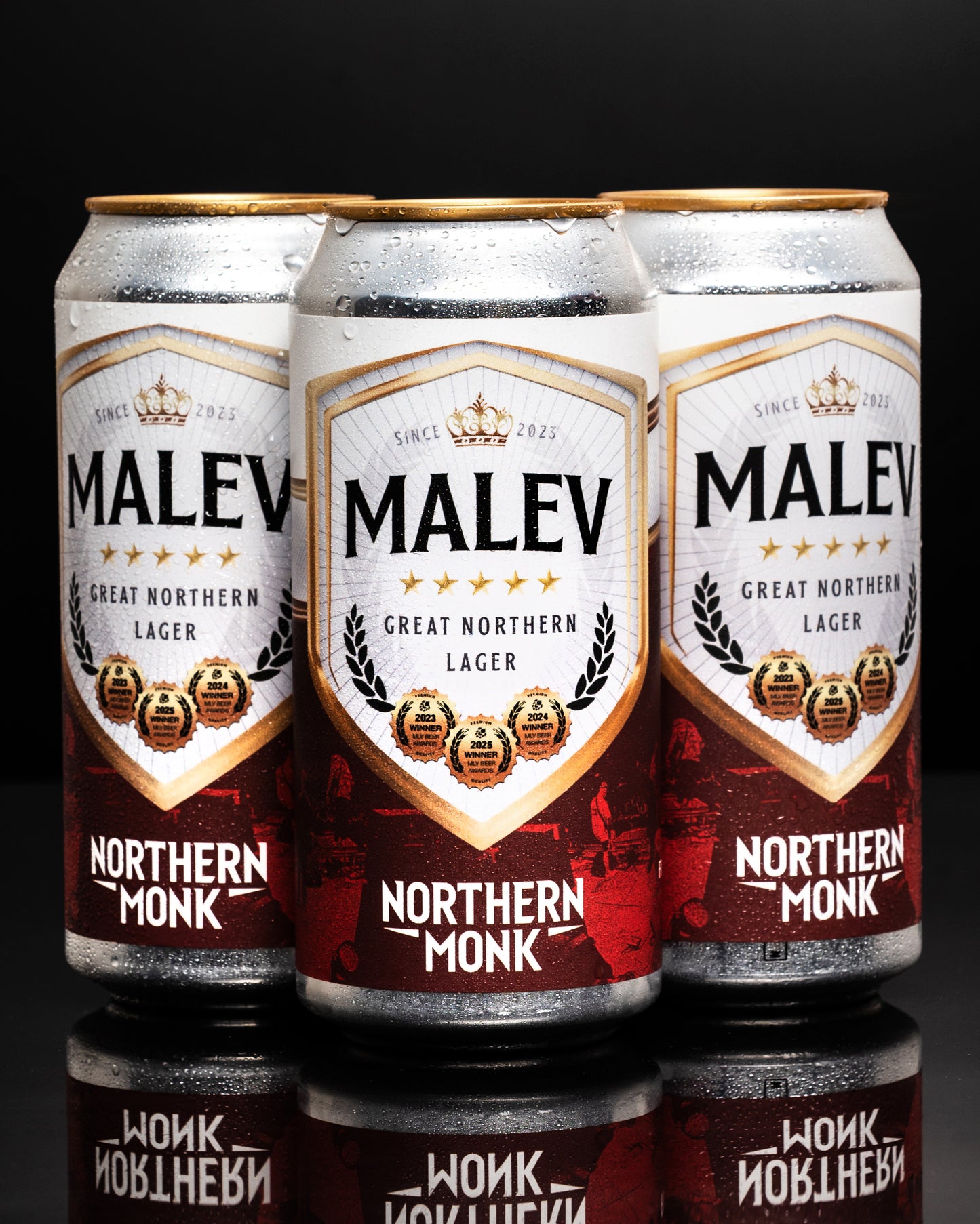 Three cans of MALEV Northern Monk Lager on a reflective surface with a dark background