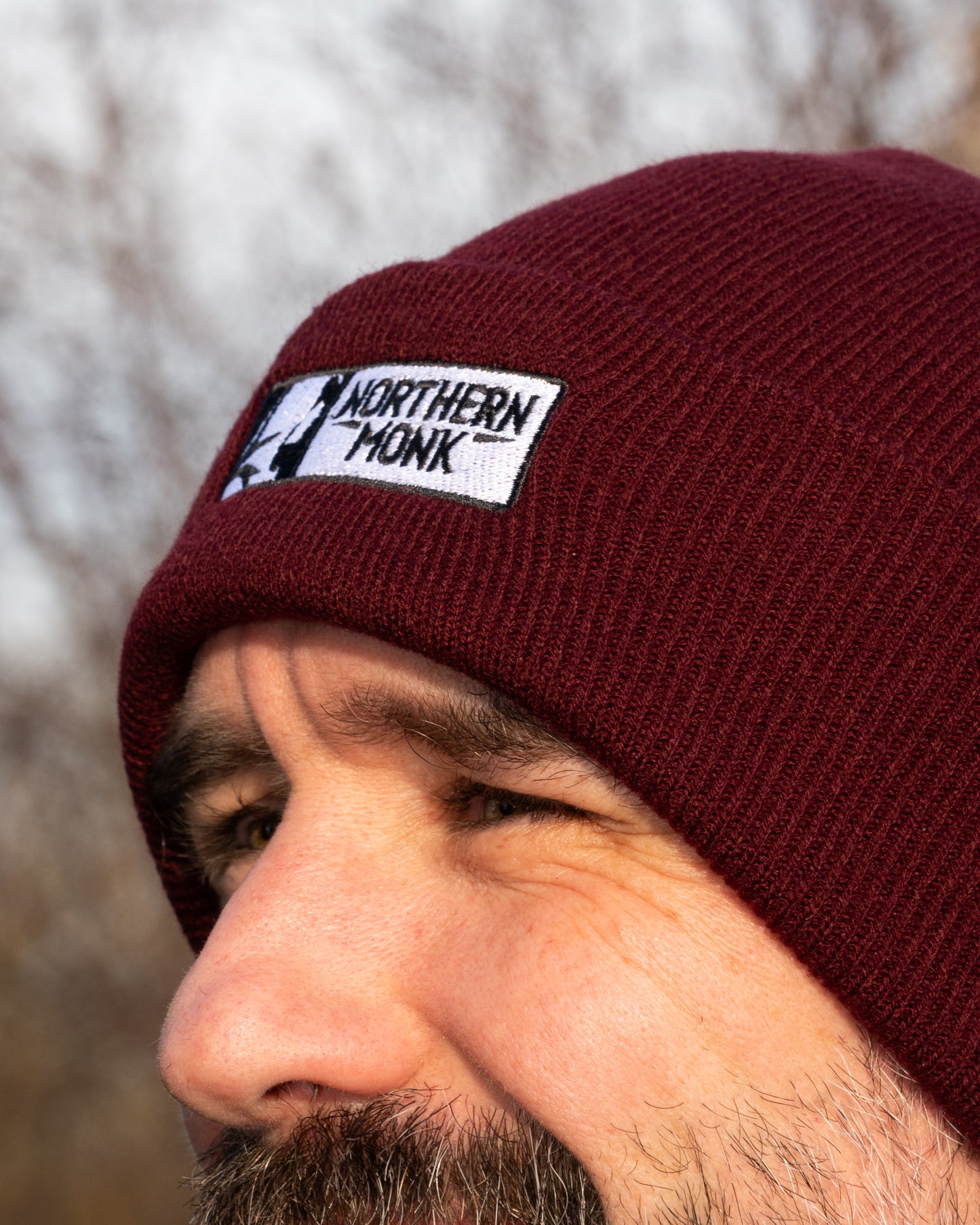 Person wearing a burgundy beanie with a 'Northern Monk' logo against a blurred natural background